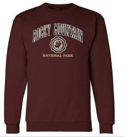 A maroon crewneck sweatshirt with a 'Rocky Mountain National Park' collegiate-style graphic on the chest, shown below a matching flat graphic print preview. The graphic features white and black lettering with a central seal containing a bison.